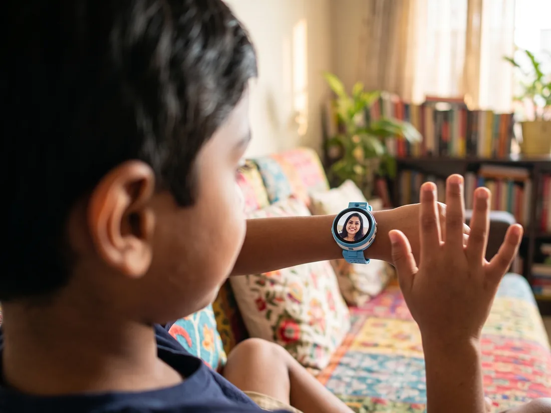 Over a boy's shoulder on the sofa at home: his sky-blue watch shows his mother on a video call as he waves to her